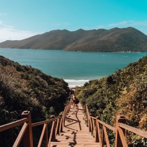 A wooden bridge surrounded by the sea and hills covered in greenery under a blue sky in Brazil
